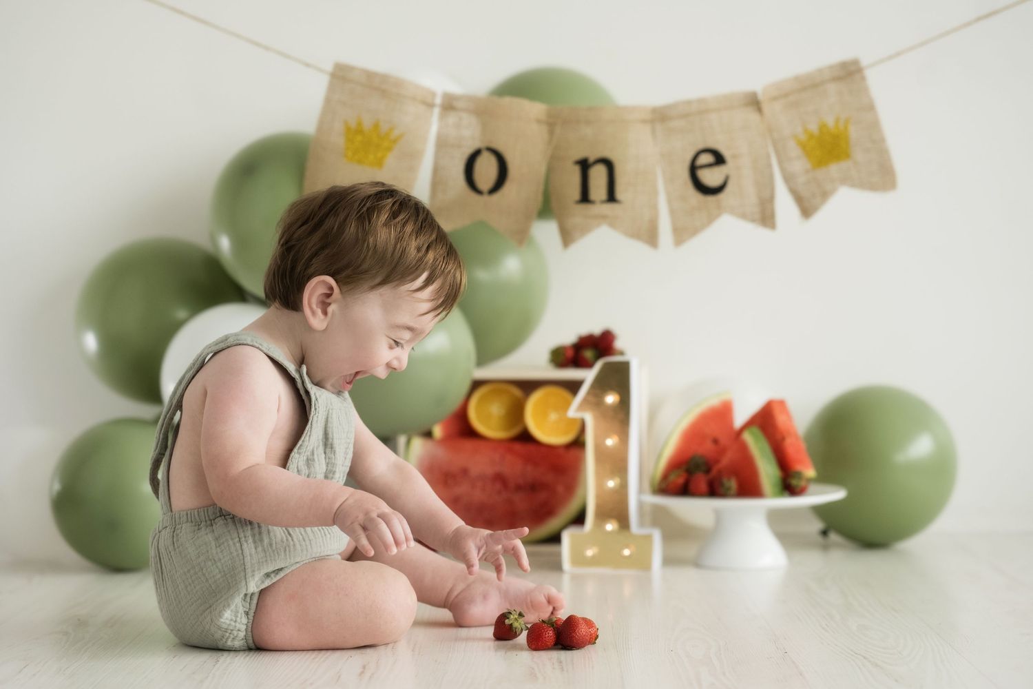 First birthday setup with green balloons, fruit display, and rustic number one decoration.