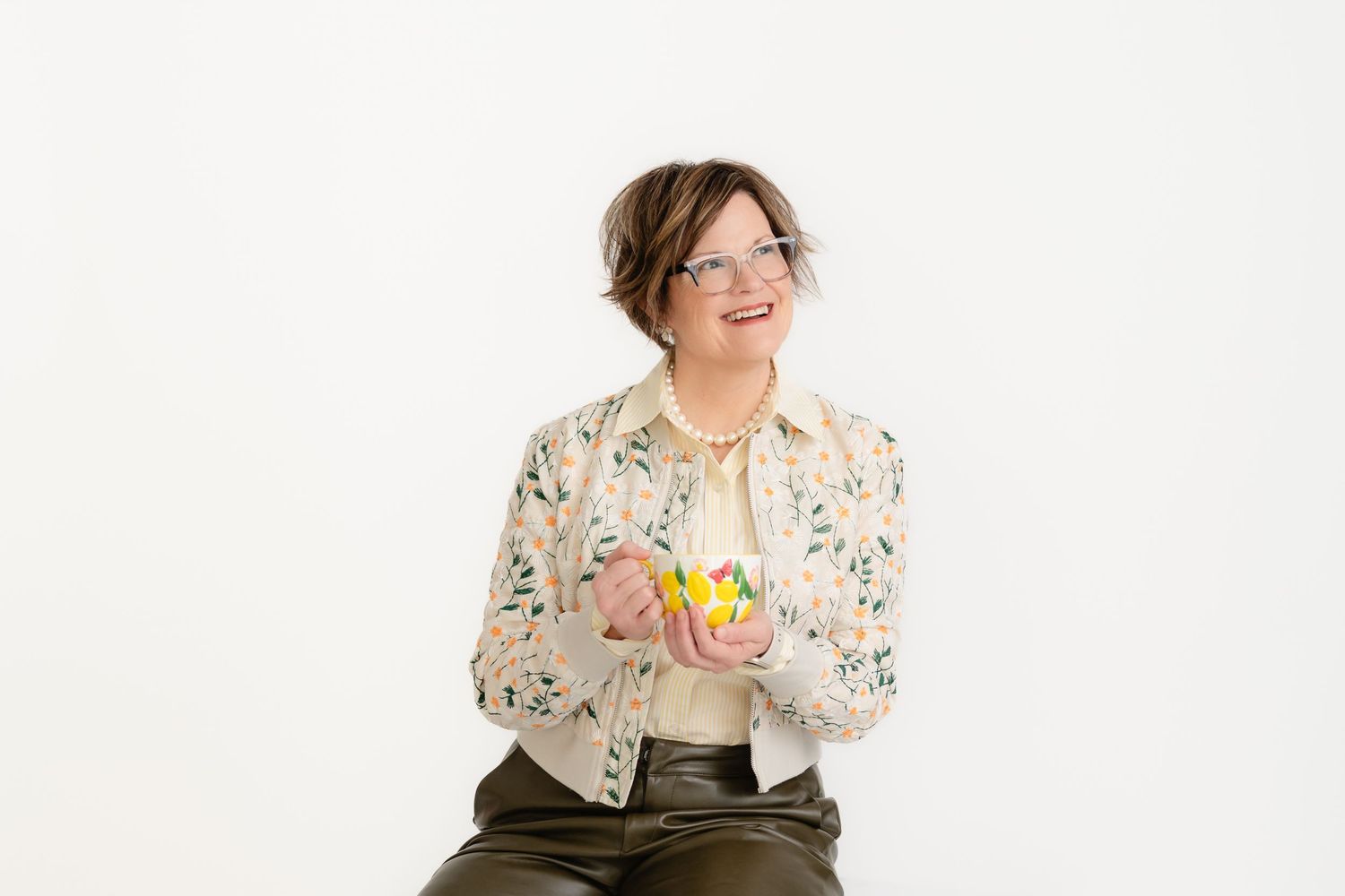 Wellness coach wearing a floral print shirt and olive pants sits against white background holding colorful flowers at Spring TX studio