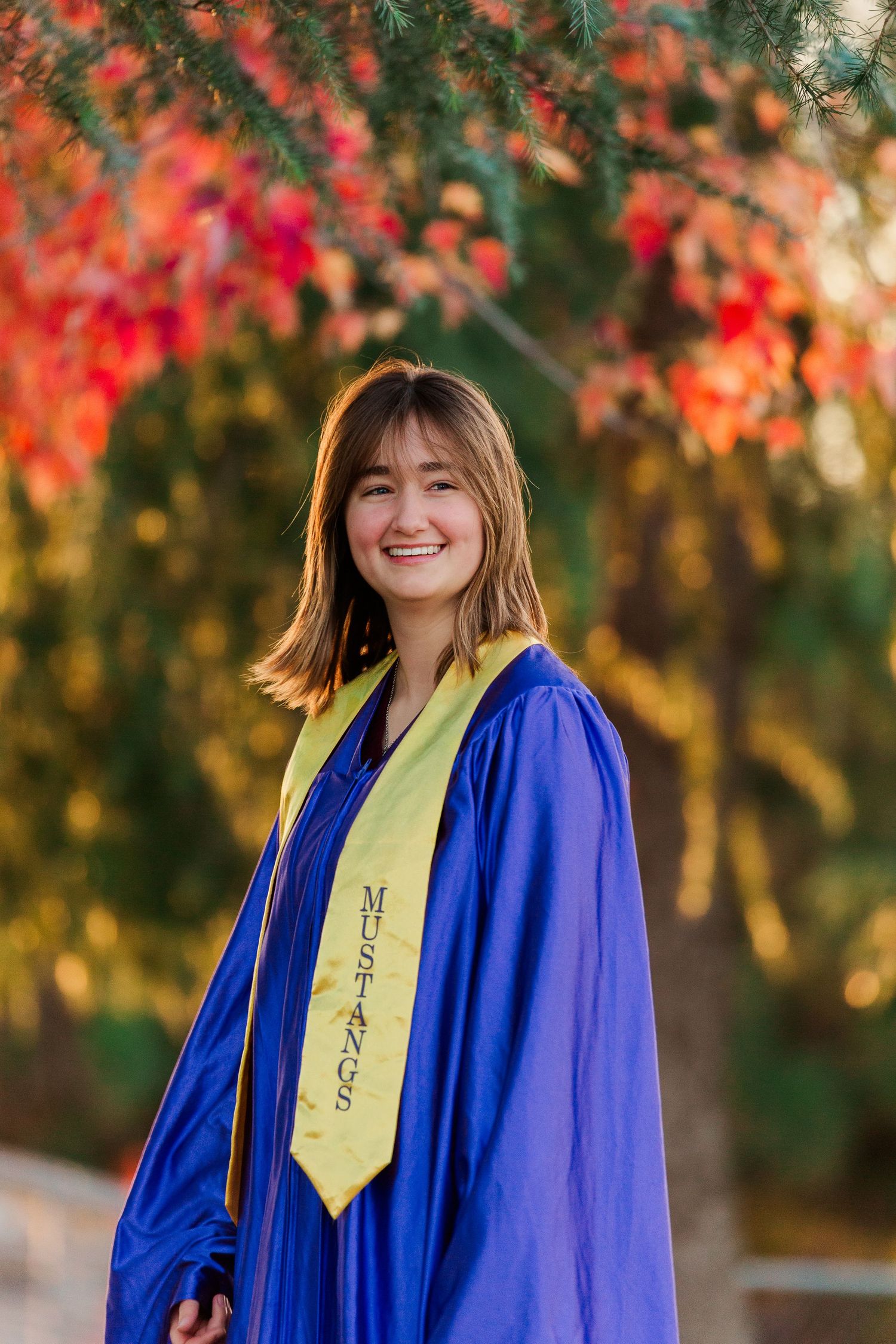 Graduate in blue cap and gown with yellow honors stole poses outdoors against vibrant autumn foliage backdrop.