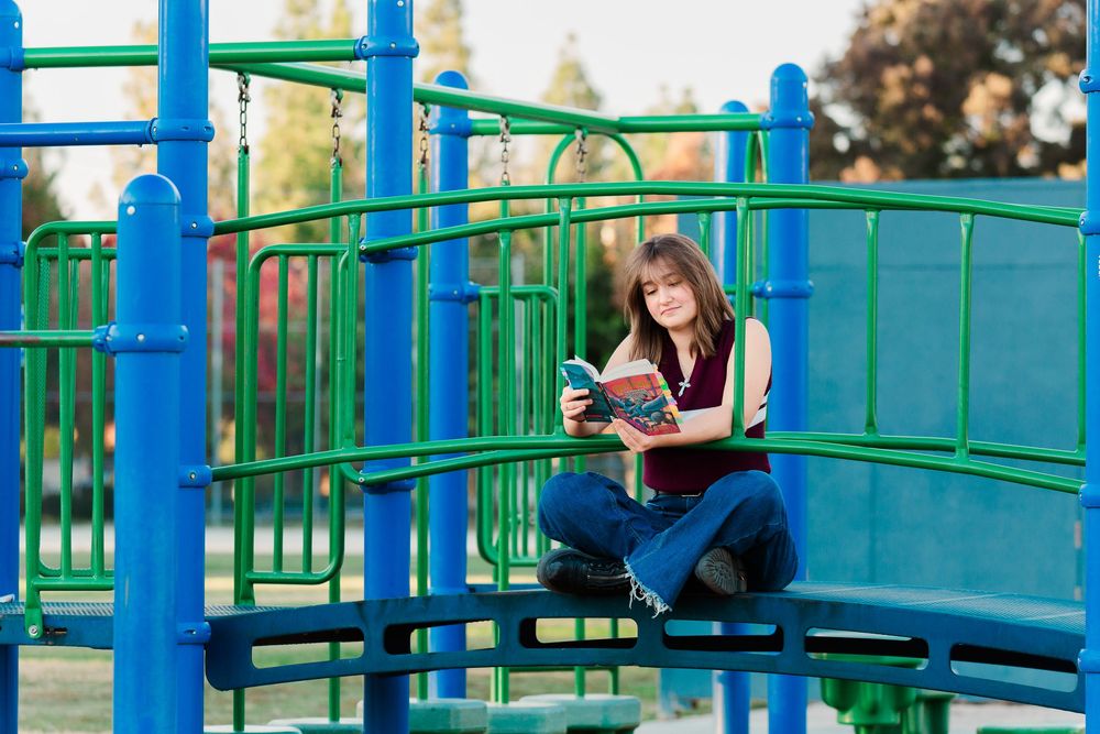 Person sits on blue and green playground equipment while reading a book in an outdoor setting.