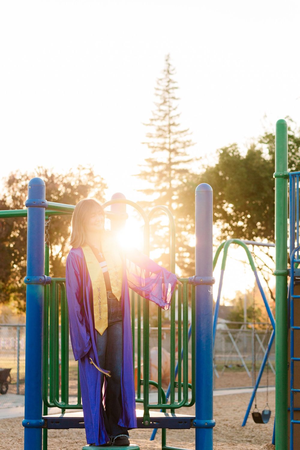 Graduate in blue and gold regalia standing on playground equipment at sunset with sunburst behind them.