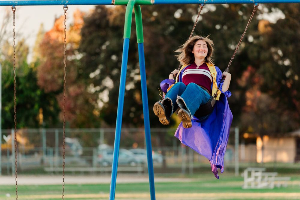 Person in a blue graduation gown swings joyfully on playground swings during sunset.