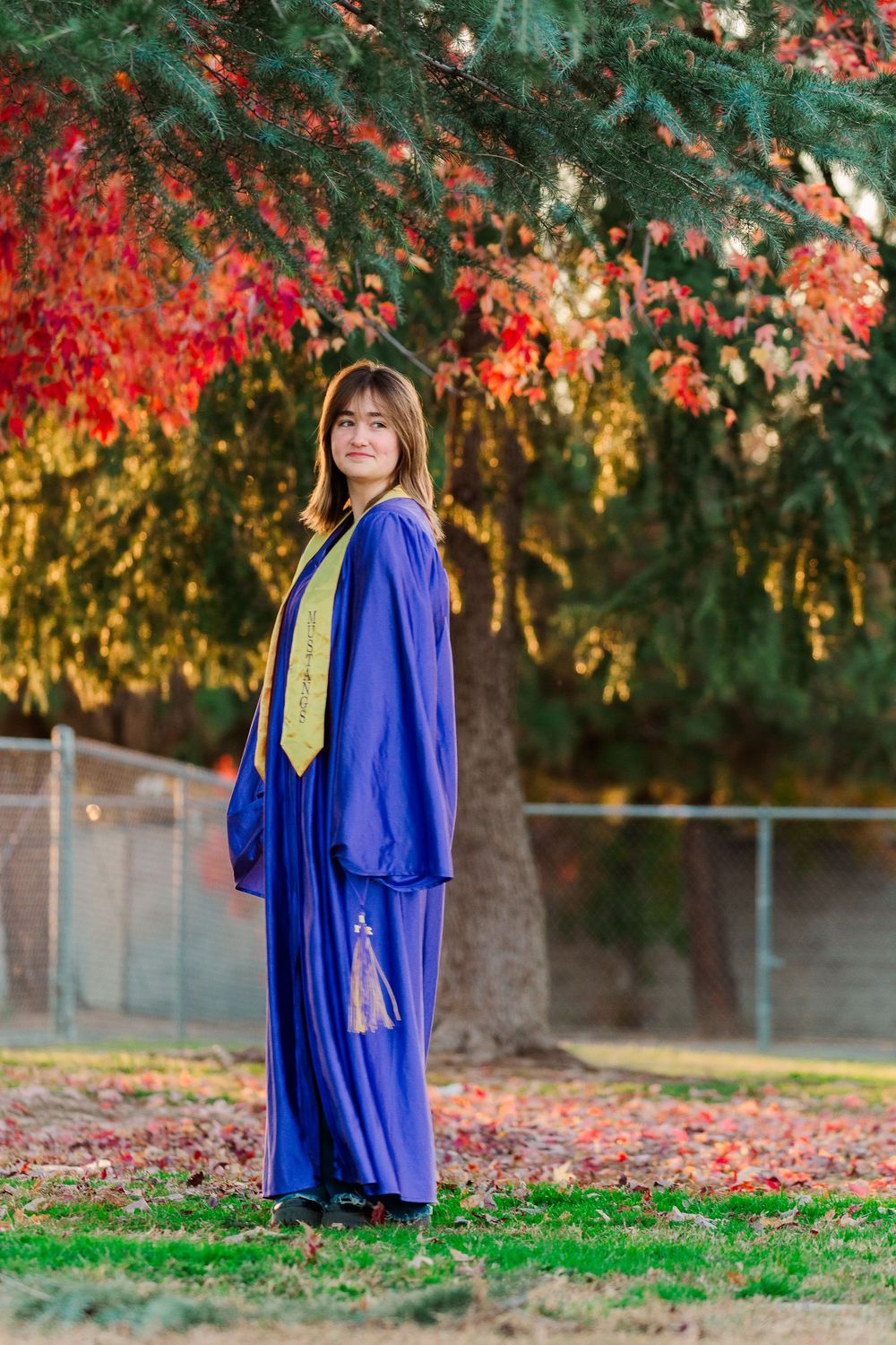 Graduate in royal blue regalia poses in front of vibrant autumn foliage with red and gold leaves.
