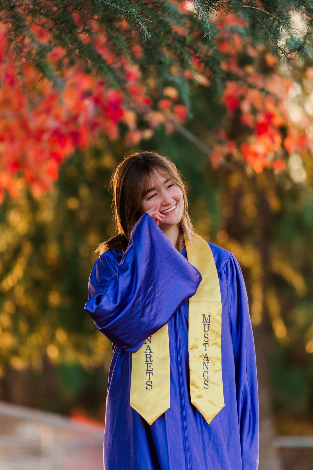 Graduate in royal blue gown with gold stoles poses under vibrant red autumn trees.