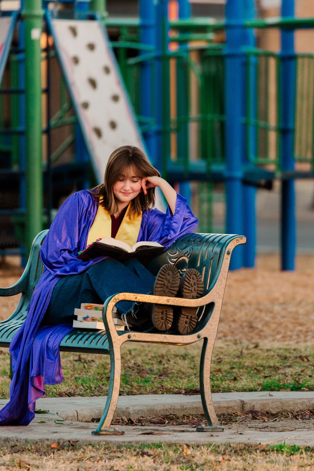 Graduate in blue cap and gown reading a book while relaxing on a park bench near playground equipment.