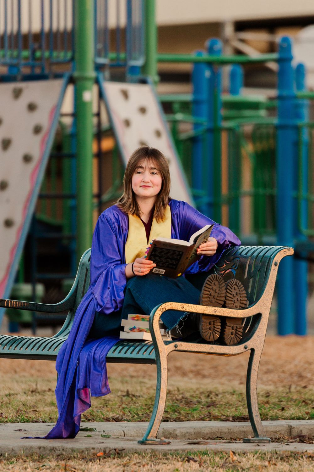 Someone relaxes on a park bench reading a book while wearing a blue graduation gown in an outdoor setting.