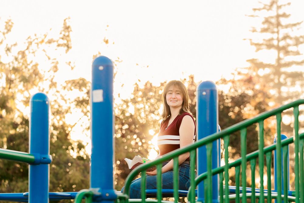 A person sits on colorful playground equipment at sunset with trees silhouetted in the background.