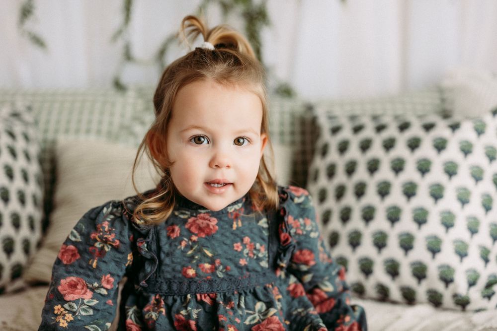 A toddler in a floral dress sits on a couch with decorative pillows in a bright living room setting.