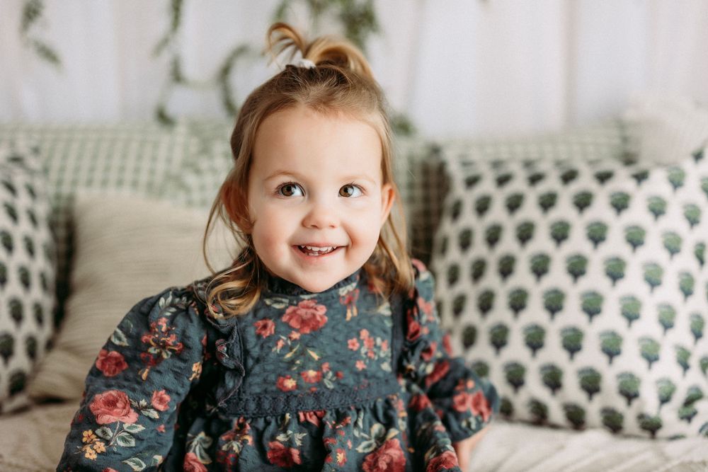 A smiling child wearing a navy floral dress sits on a couch with patterned pillows in a bright room.
