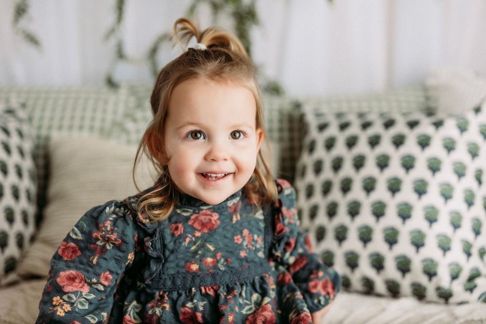 A toddler in a floral dress smiles and plays while sitting on a patterned couch.