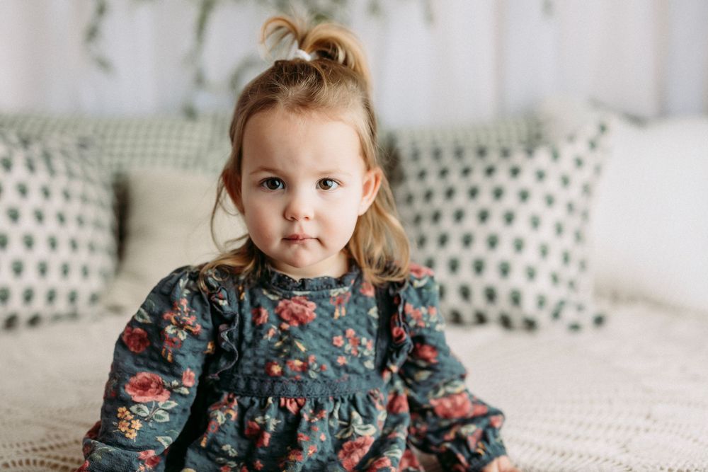 A child wearing a floral dress sits on a white textured surface in a bright, airy setting.