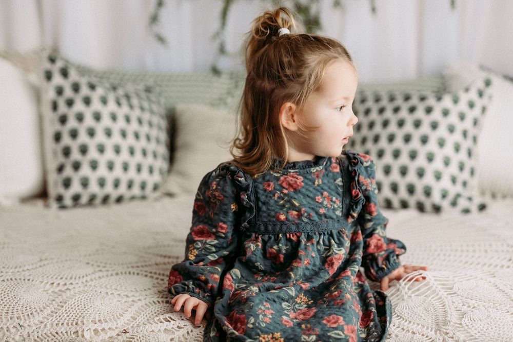 A child in a floral dress sits and plays on a white couch with polka dot pillows in a bright room.