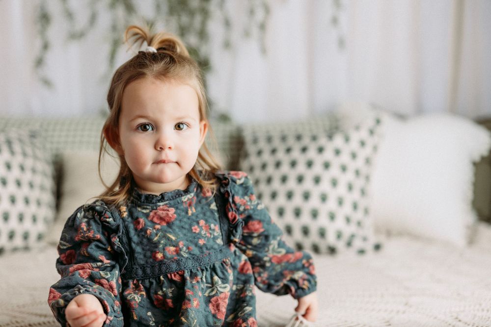 A baby wearing a floral dress sits on a white couch with polka dot pillows in the background.