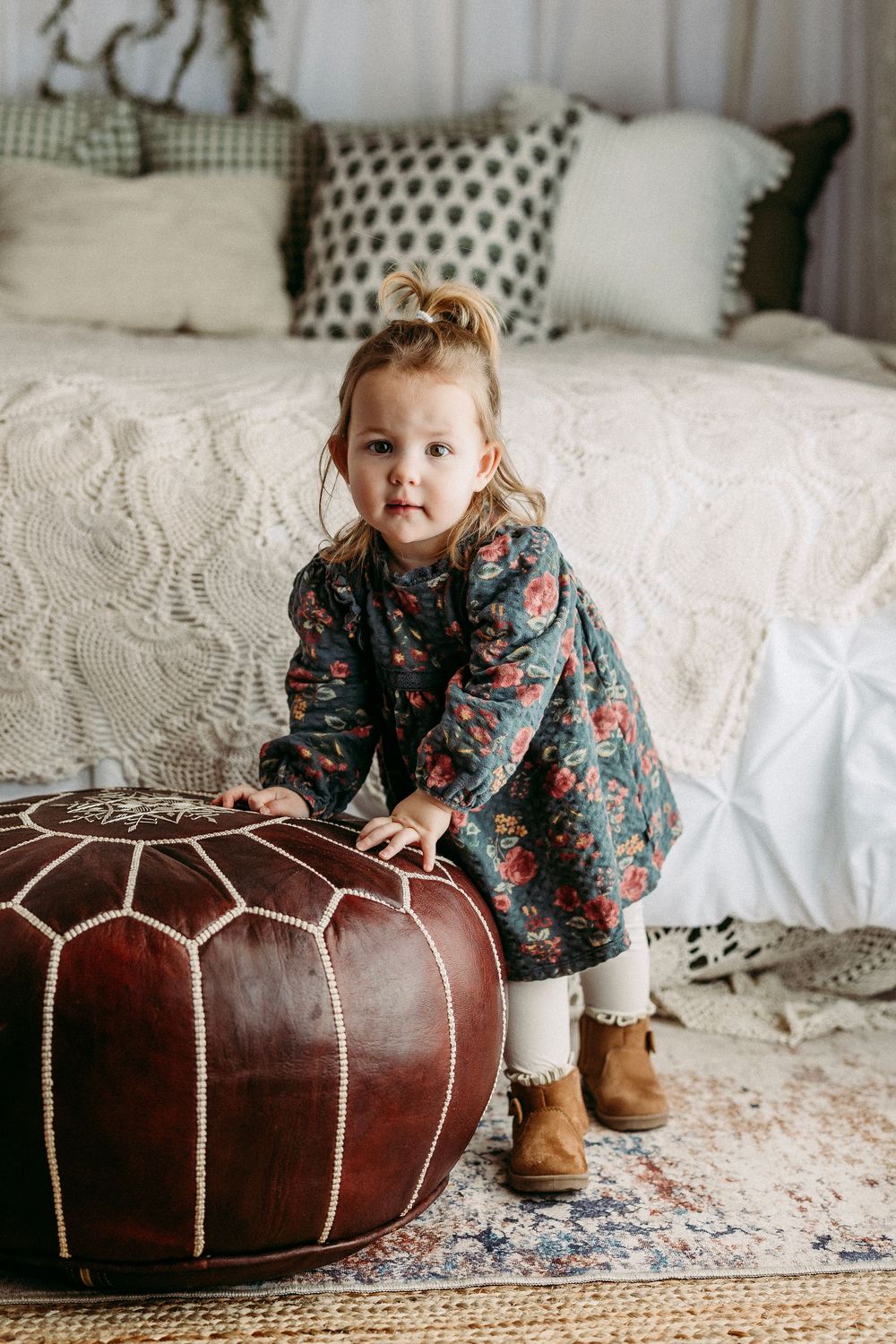 Small child in floral dress plays on white bedding near brown leather poufs in cozy bedroom setting.