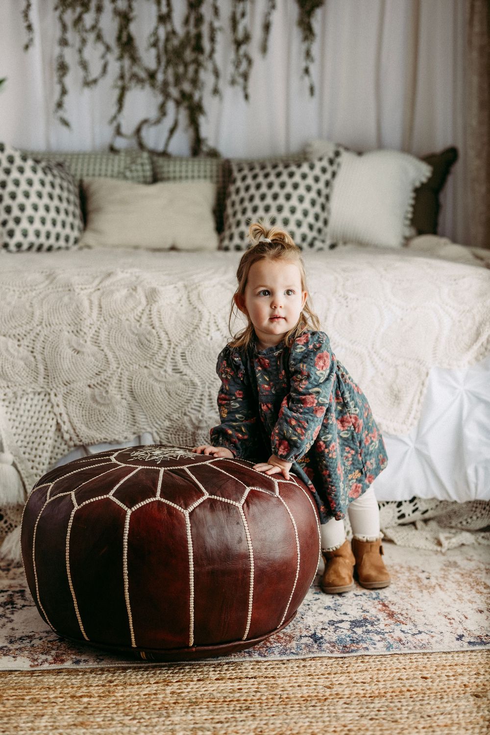 A child sits beside a brown leather pouf on a white textured bedspread with decorative pillows and natural jute rug.