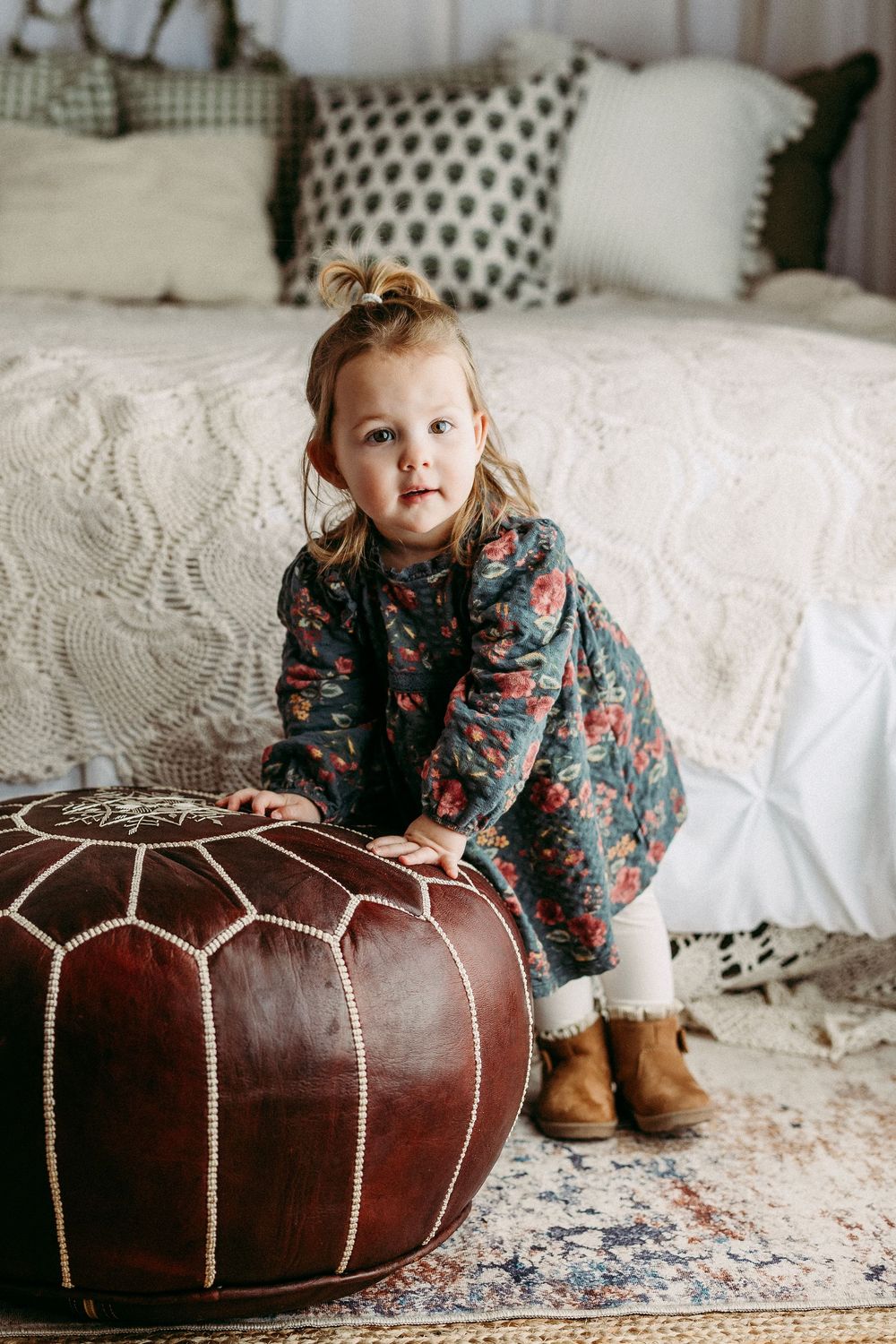 A child wearing a floral print top plays near a brown leather pouf ottoman on a patterned rug.