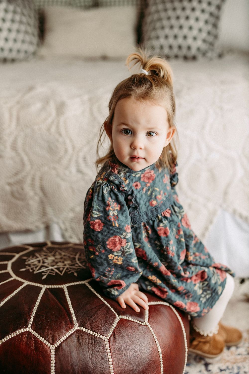 A small child in a floral dress sits on a brown leather ottoman with decorative white stitching.