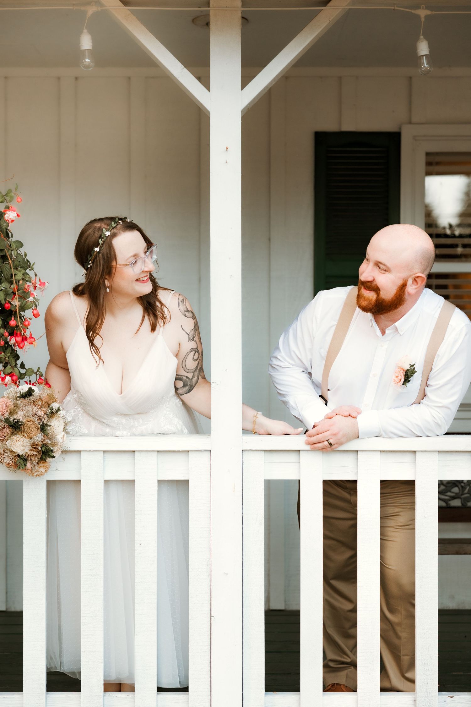 A couple in white wedding attire shares a romantic moment while leaning on a white porch railing. Metro Detroit Wedding Content Creator