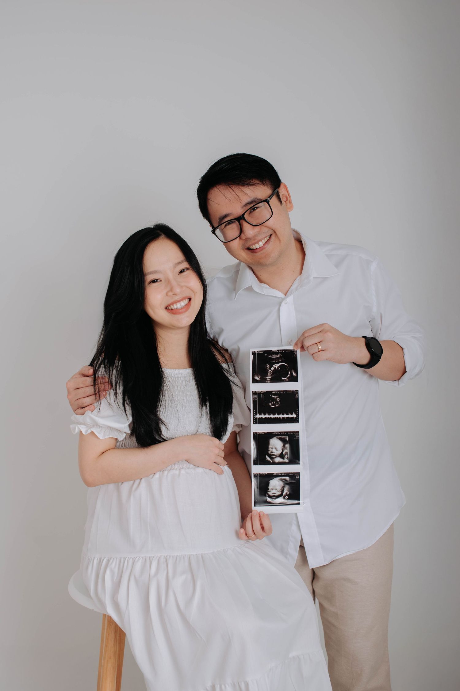 An expectant mother poses in a flowing off-white tiered maxi dress against a neutral backdrop during a maternity photoshoot.