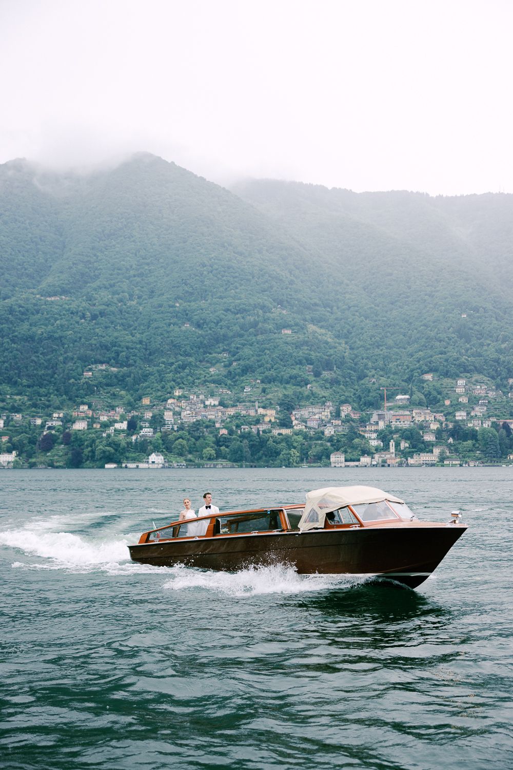 Classic wooden boats speed across a misty lake with mountains and coastal homes in the background.