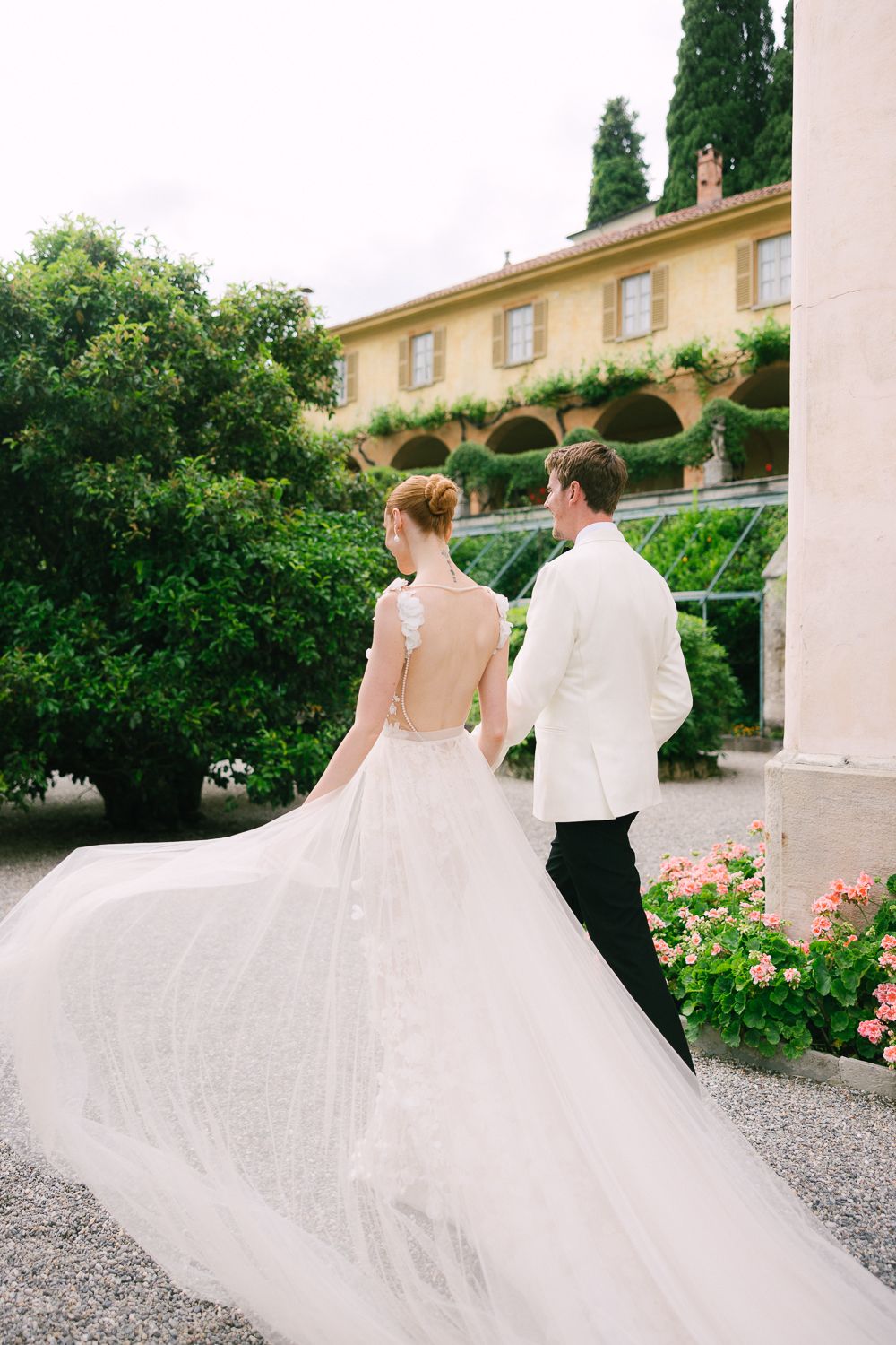 A romantic wedding couple walks through a garden path with lush greenery and blooming roses at an elegant venue.