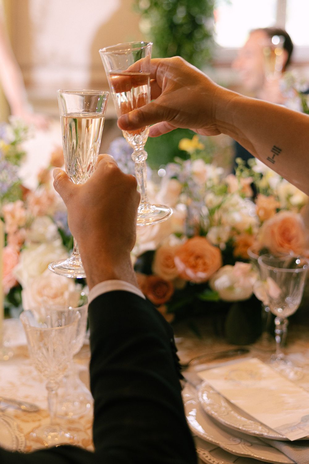 Two hands raise crystal champagne flutes for a toast at an elegant table with peach roses and floral centerpieces.