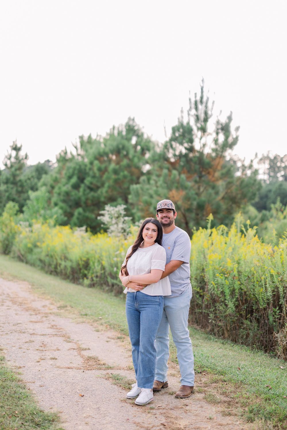Romantic couple embracing on a rustic dirt path with yellow flowers and pine trees in the background.