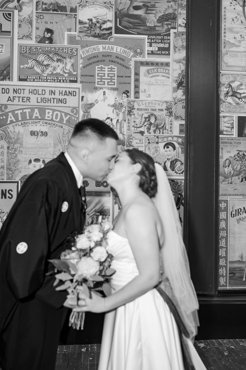 A black and white wedding photograph shows a romantic kiss against a wall covered in vintage posters and advertisements.