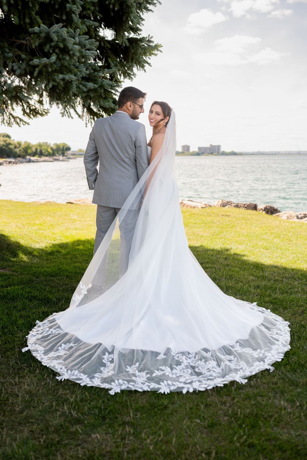 A couple shares an intimate moment on a waterfront lawn, with the bride wearing a stunning lace-trimmed cathedral veil.