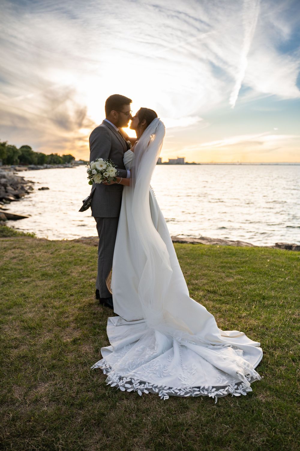 A romantic sunset wedding photo series showing a couple embracing on a grassy oceanfront with flowing white wedding dress details.