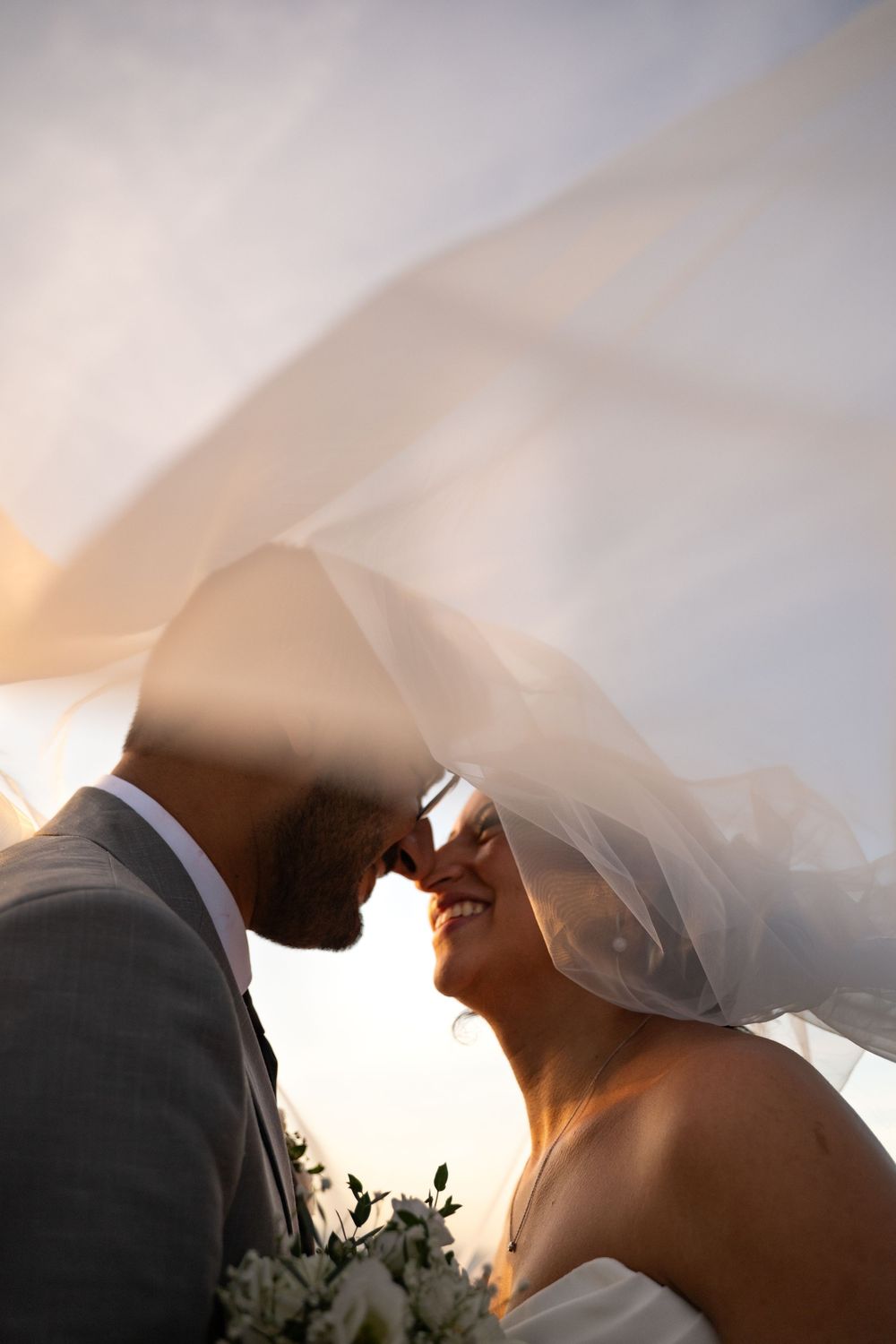 A romantic silhouette of a couple under a flowing white wedding veil during sunset.