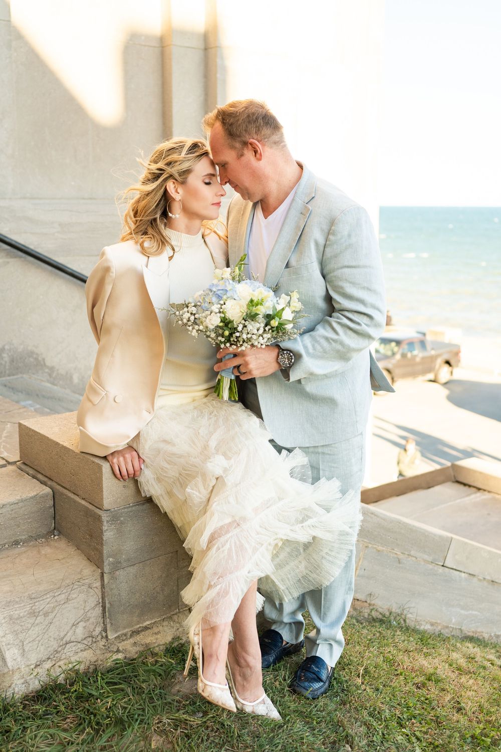 A romantic beach wedding scene with a couple sharing an intimate moment on stone steps while holding a white bouquet.