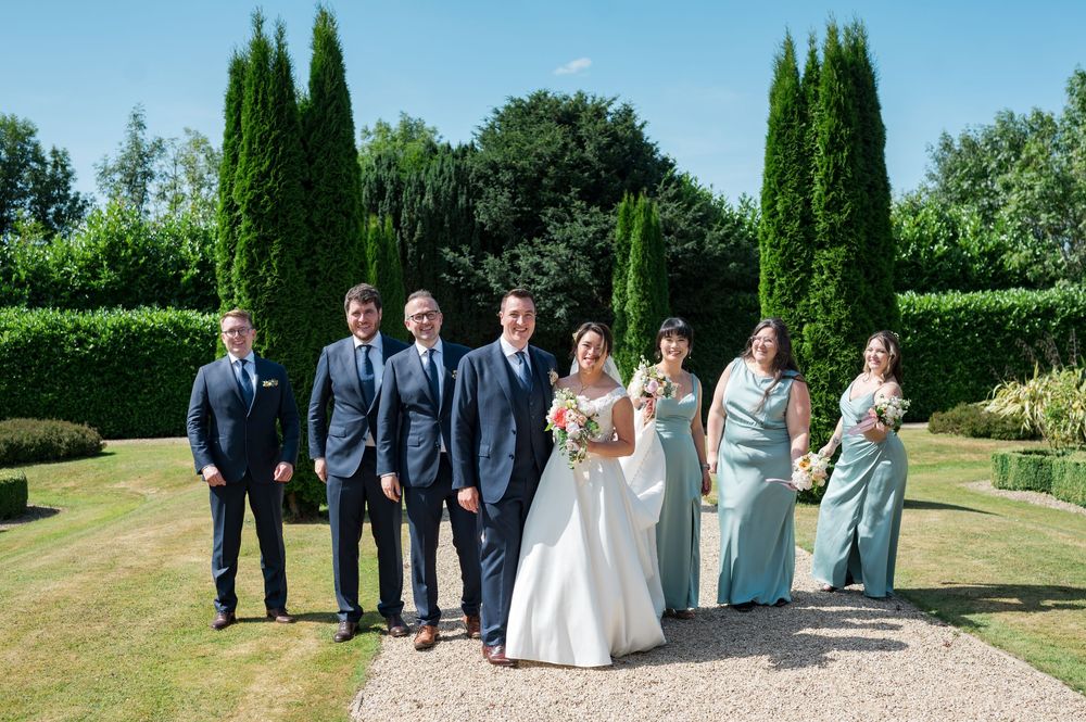 Wedding party poses on manicured lawn with tall cypress trees and hedges in elegant garden setting.