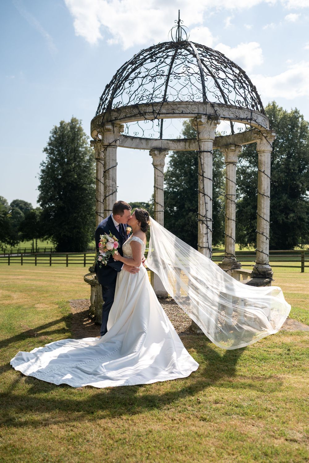 A romantic wedding scene under an ornate metal gazebo with a flowing white veil and dress train on a sunny day.
