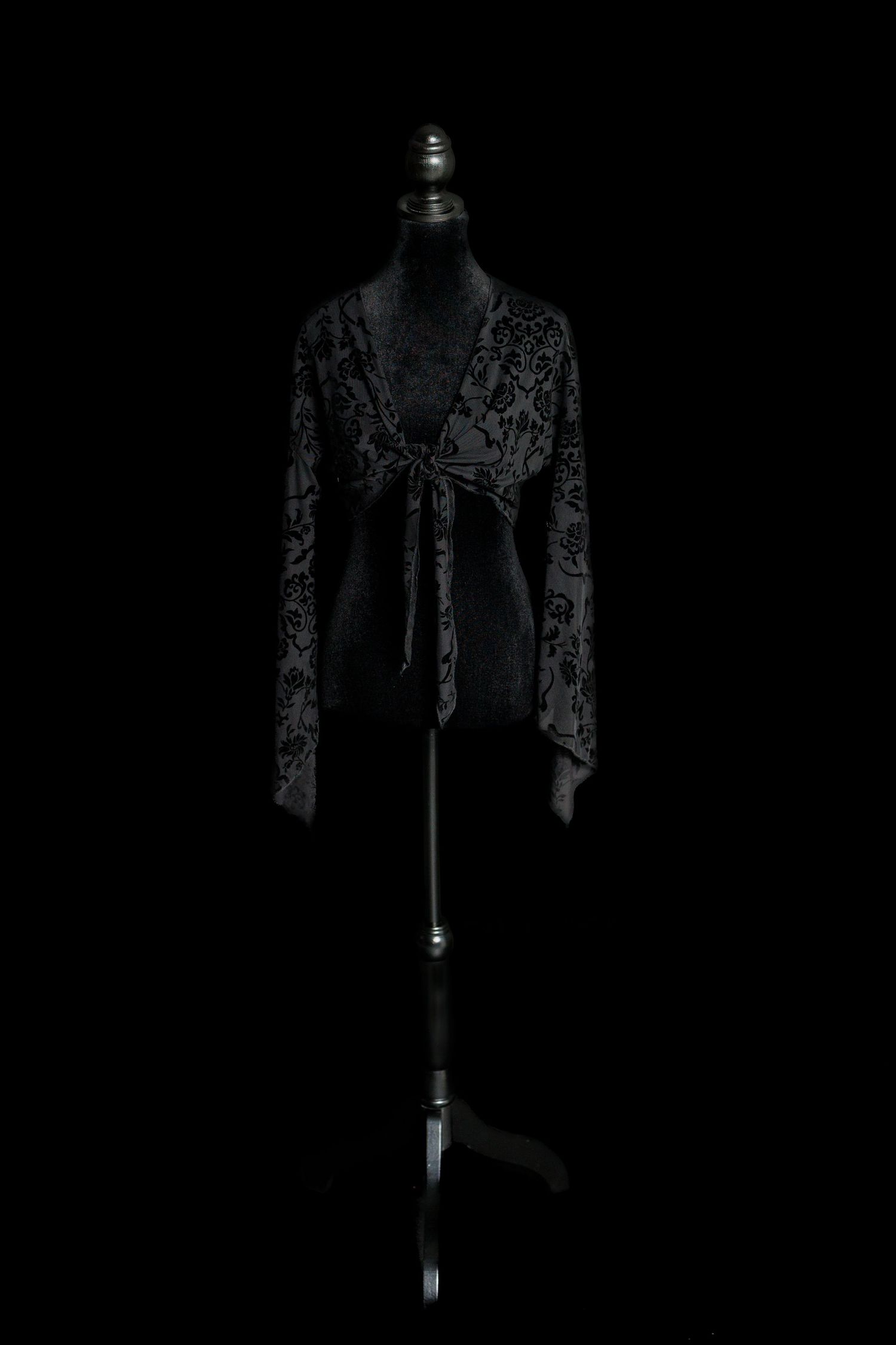 Black sheer maternity top with velvet floral pattern, tie-front closure, deep V neckline, and long flared sleeves on a dress form.