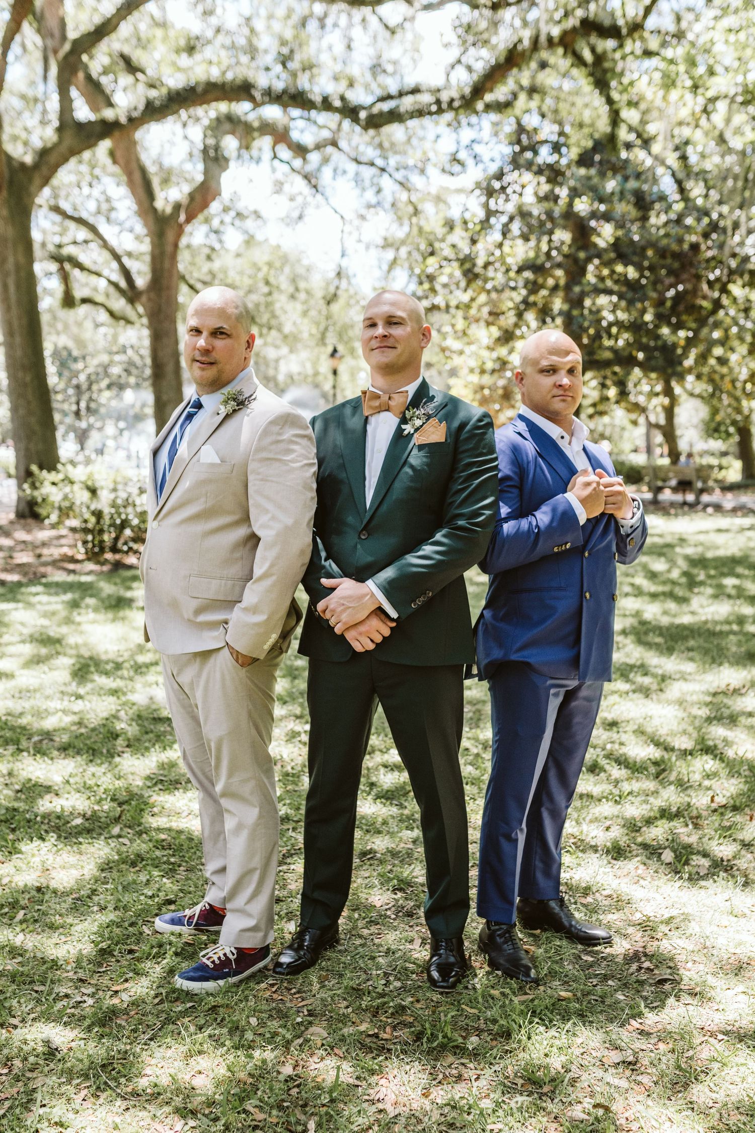 Wedding party members in suits pose together on sunny lawn under trees.