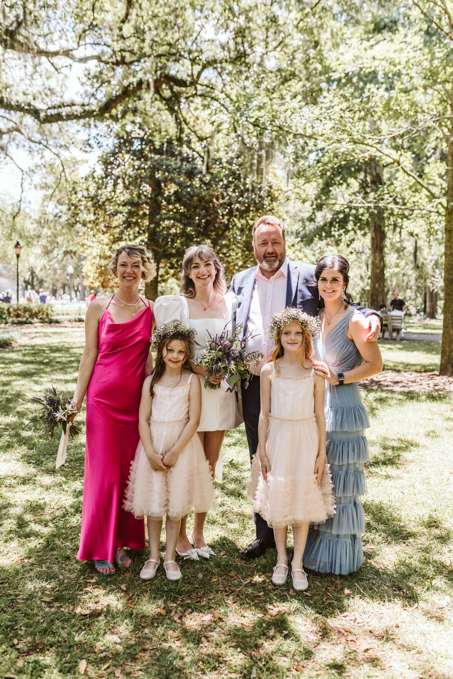 Wedding party poses under shaded trees in a garden setting wearing coordinated formal attire.
