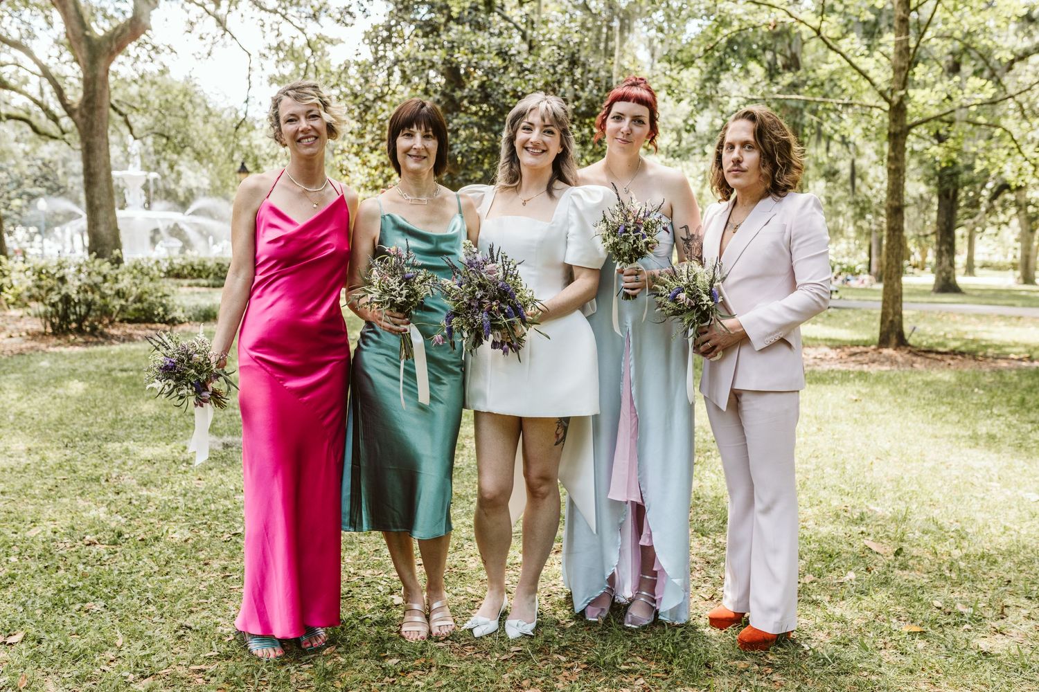 Wedding party in colorful dresses poses together outdoors in a sunny garden setting.