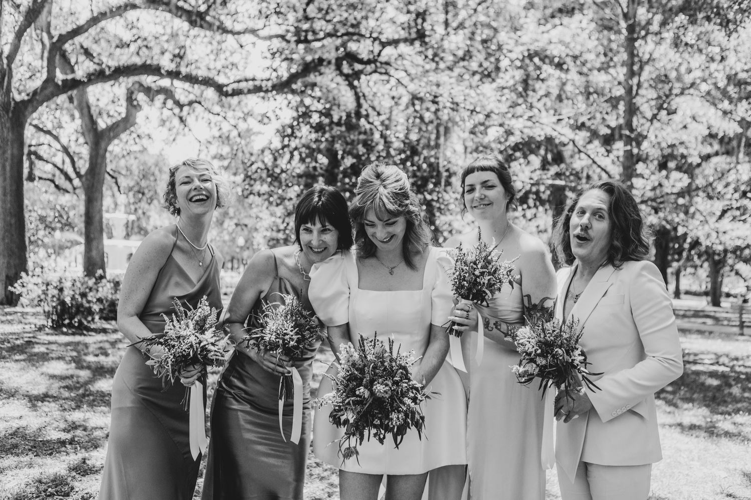 Wedding party laughing together under trees draped with Spanish moss in an outdoor garden setting.