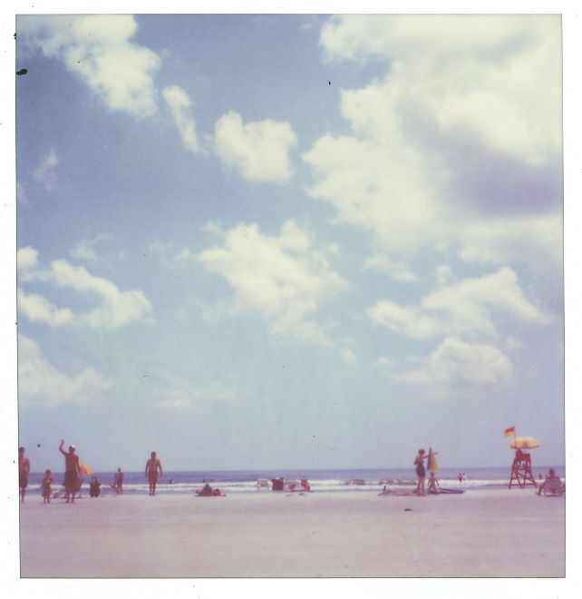 Beachgoers enjoying a sunny day on a sandy shore with lifeguard towers visible in the distance under puffy clouds.