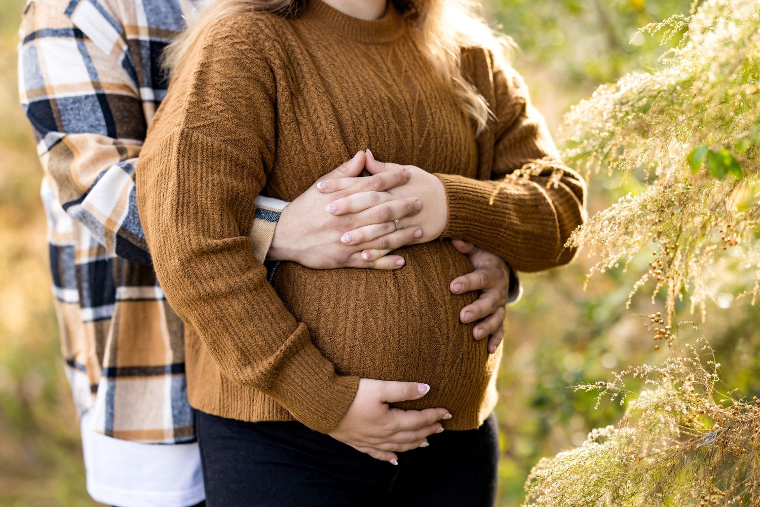 Close up view of hands cradling a pregnant belly while wearing a cozy brown knit sweater in outdoor autumn setting.