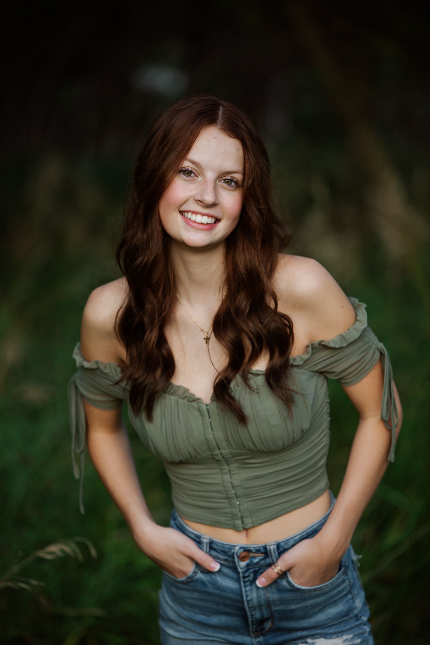 A person with long brown hair wearing an olive green off-shoulder top and blue jeans poses outdoors with a joyful expression.