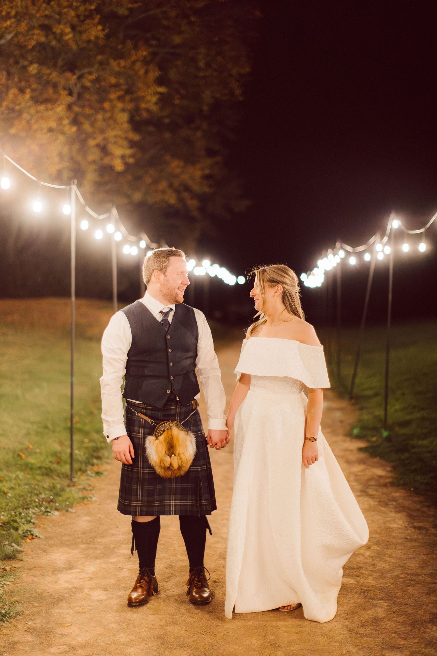 A romantic evening wedding scene with string lights illuminating a path where a couple walks together in traditional Scottish attire.