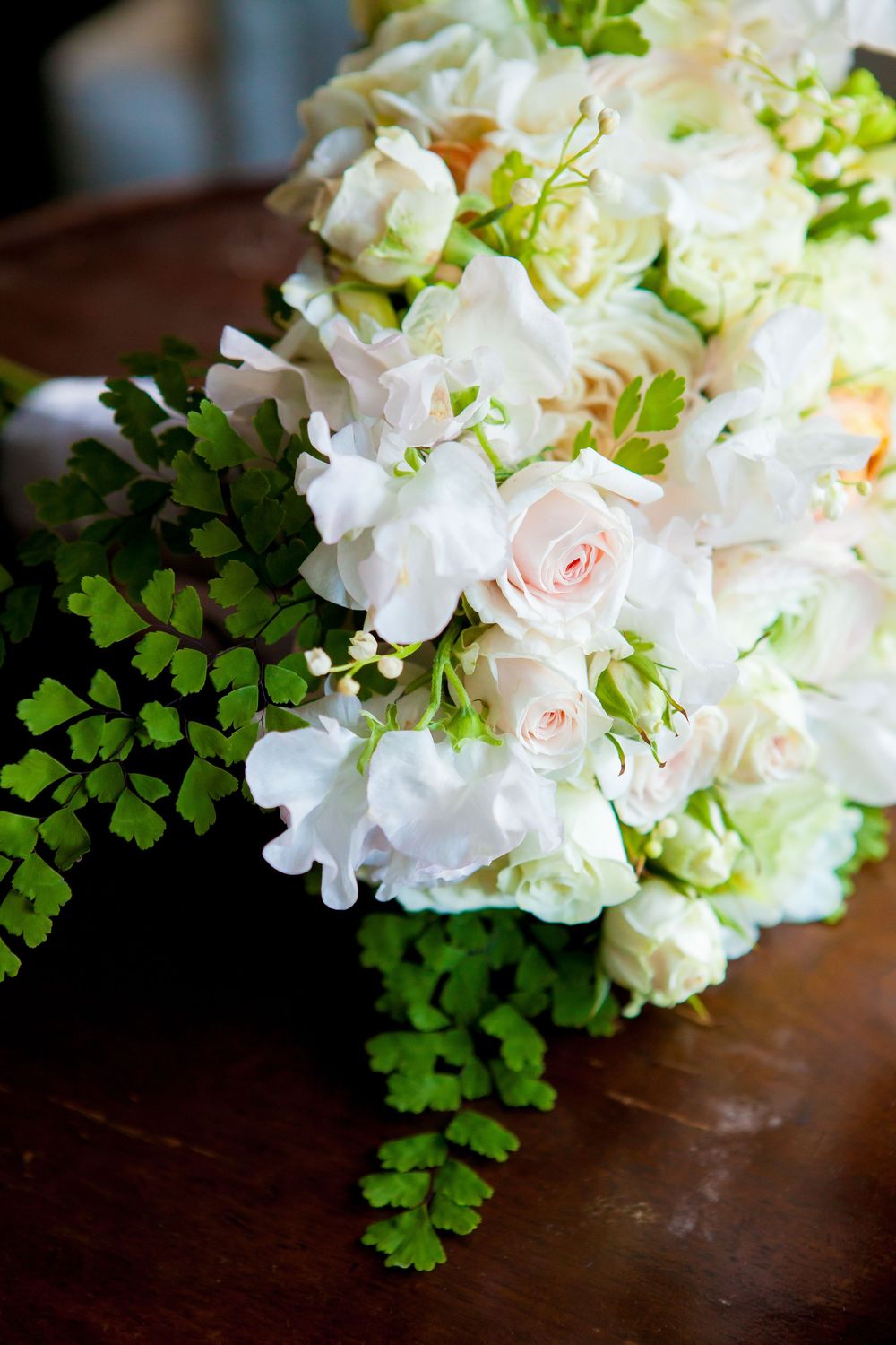 Delicate white and blush pink roses arranged with green ferns on a dark wooden surface.