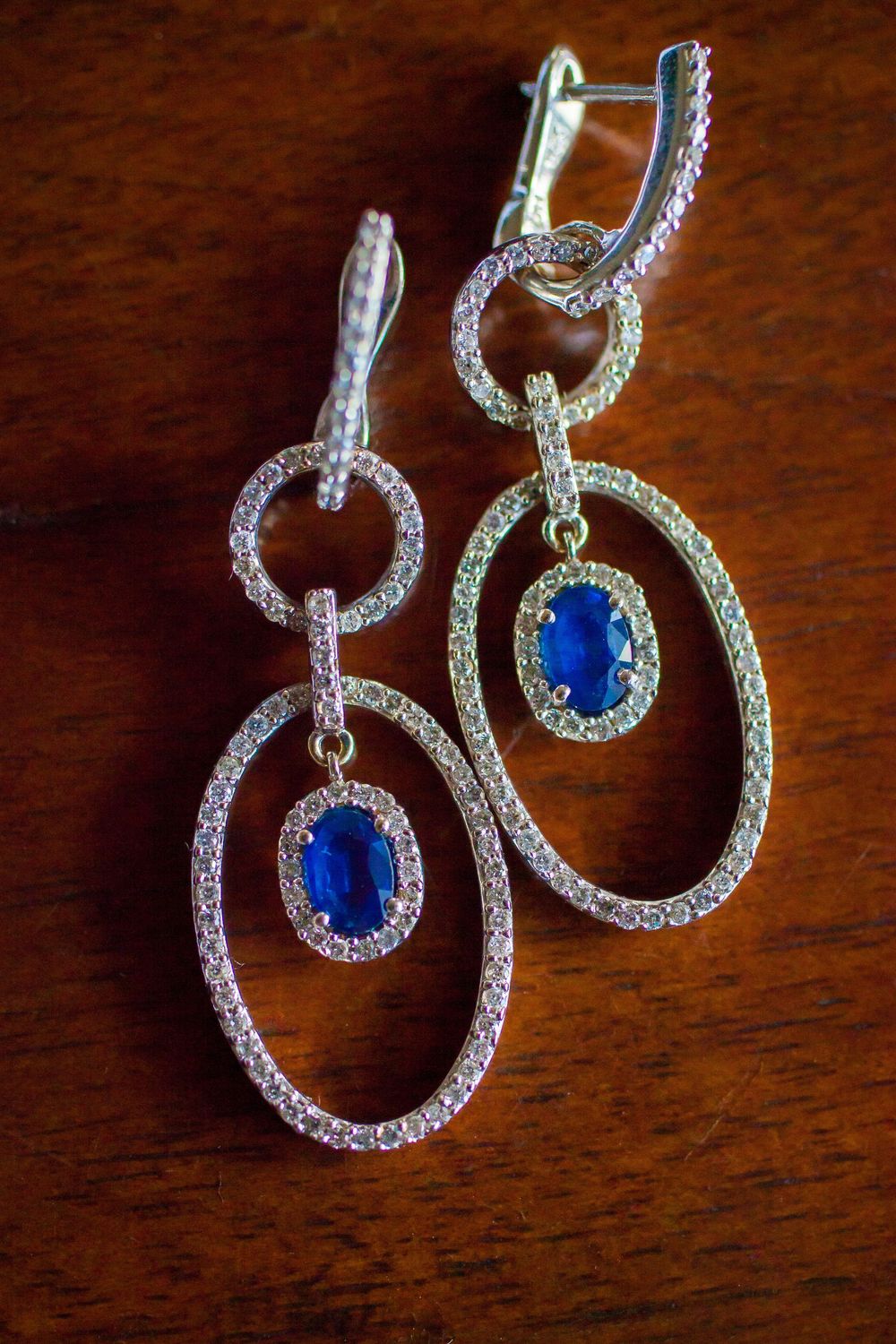 Elegant silver and sapphire drop earrings with crystal-encrusted circular design sparkle against dark wooden surface.