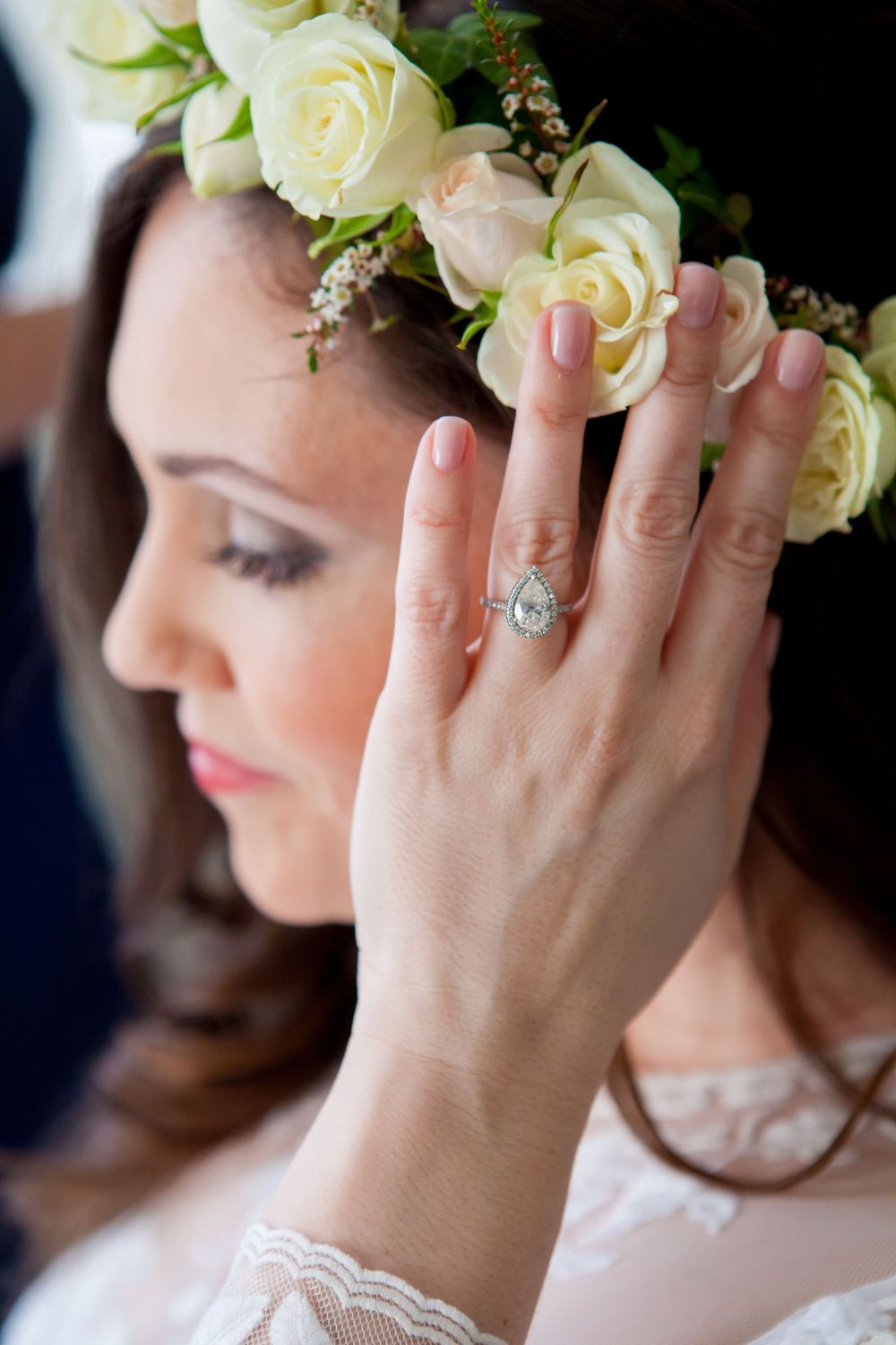 A delicate floral crown of white roses adorns the head while a sparkling engagement ring is displayed on the hand.