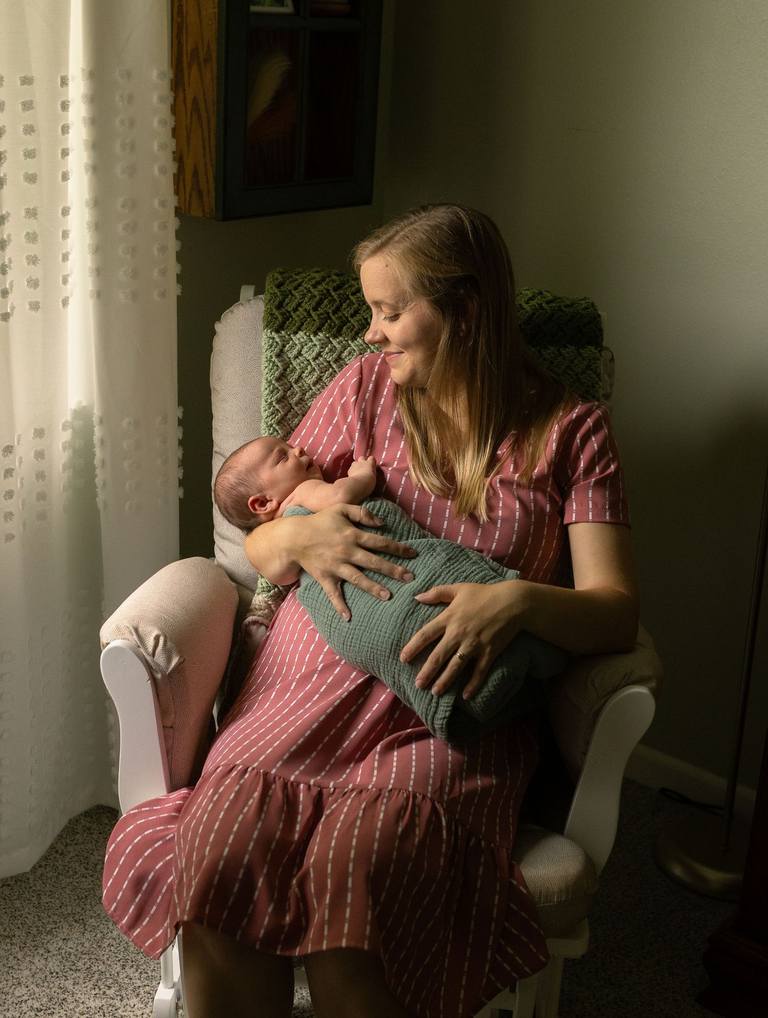 Mother in pink dress holding baby near window with soft natural lighting.