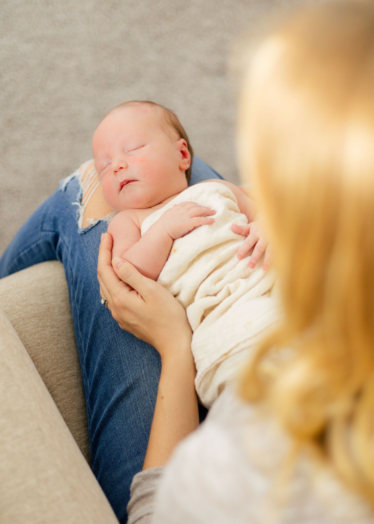 Parent cradling sleeping baby in cream outfit on grey couch.