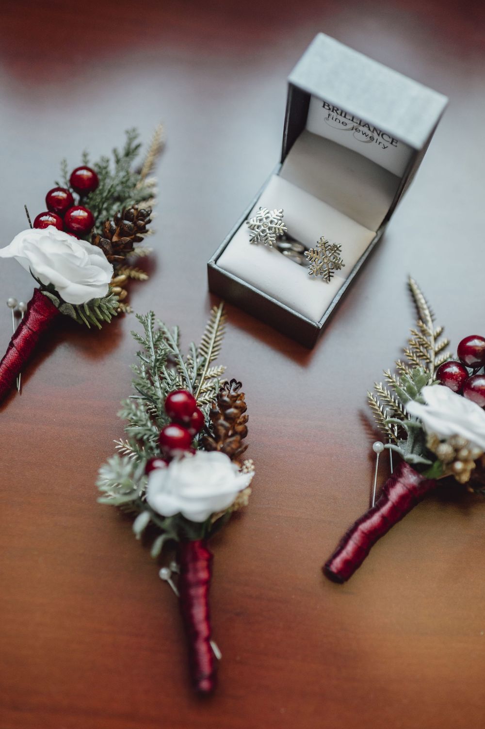 Wedding boutonniere with burgundy ribbon, pine sprigs, red berries and pinecones rests on wooden surface.