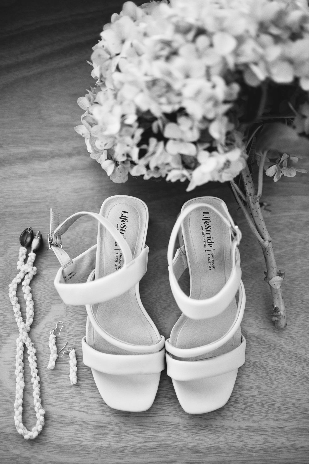 Black and white photo of white bridal sandals with pearl necklace and hydrangea flowers on wooden surface.
